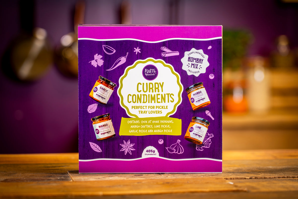 A boxed food product labeled 'Curry Condiments' stands upright on a wooden surface with a blurred kitchen background. The package is predominantly purple with a large cream/yellow central label and small illustrations of spice jars and ingredients.