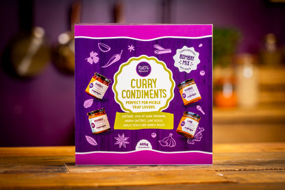 A boxed food product labeled 'Curry Condiments' stands upright on a wooden surface with a blurred kitchen background. The package is predominantly purple with a large cream/yellow central label and small illustrations of spice jars and ingredients.