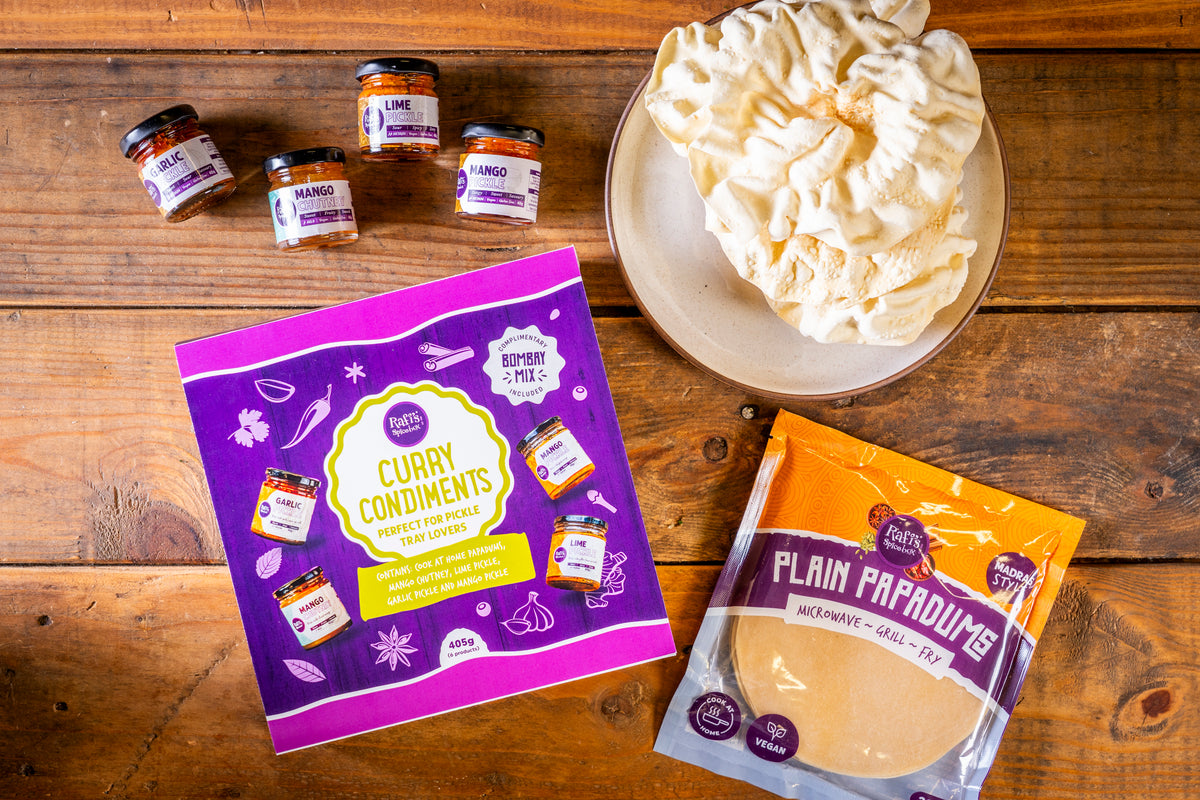 A styled flat-lay of a food product assortment on a rustic wooden table, featuring a purple box of Curry Condiments, a bag of Plain Papadums and a plate of cooked Papadums